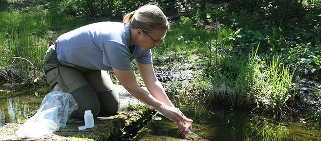 En kvinna sitter på huk vid ett vattenbryn. Hon håller i ett mätinstrument. Foto.