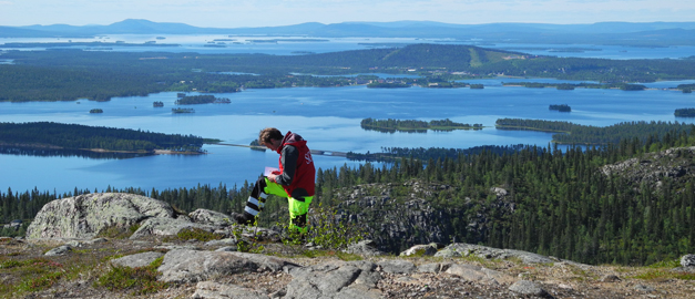 En man står och antecknar i ett block. I bakgrunden syns vatten och fjäll. Foto.