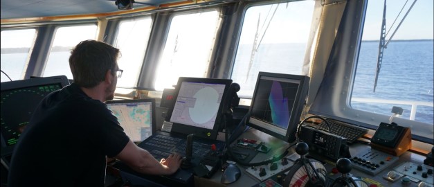 En person sitter och styr ett fartyg. Fönster ut mot hav. Styrinstrument